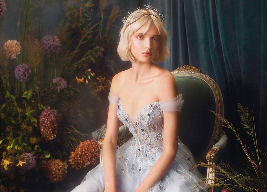 Woman in bejeweled tiara and blue off-shoulder gown sits in a vintage chair.