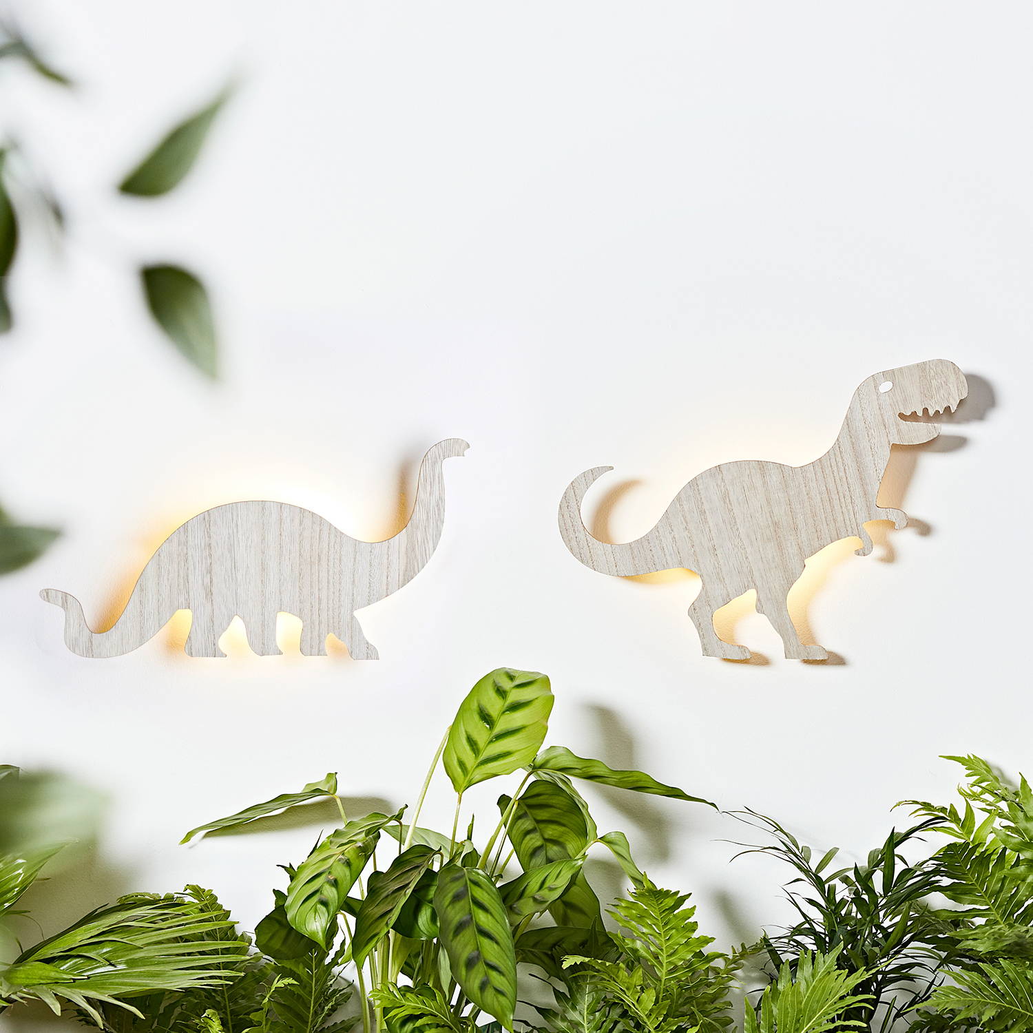 2 Wooden dinosaur wall lights against a white wall with plants and foliage around them.