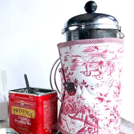 a handmade french press cozy with a button on a french press with a box of tea next to it