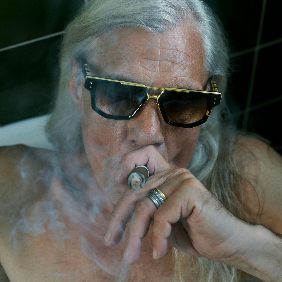 Elderly man smoking a cigar and wearing sunglasses, surrounded by smoke.
