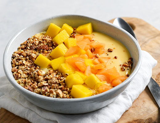 Bowl of smoothie topped with mango, granola, and peach slices.