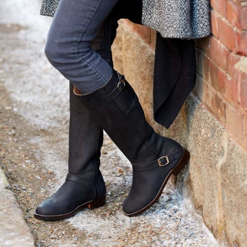 Woman leaning against a brick wall wearing black leather Reina wide calf equestrian boots with brass buckle details.