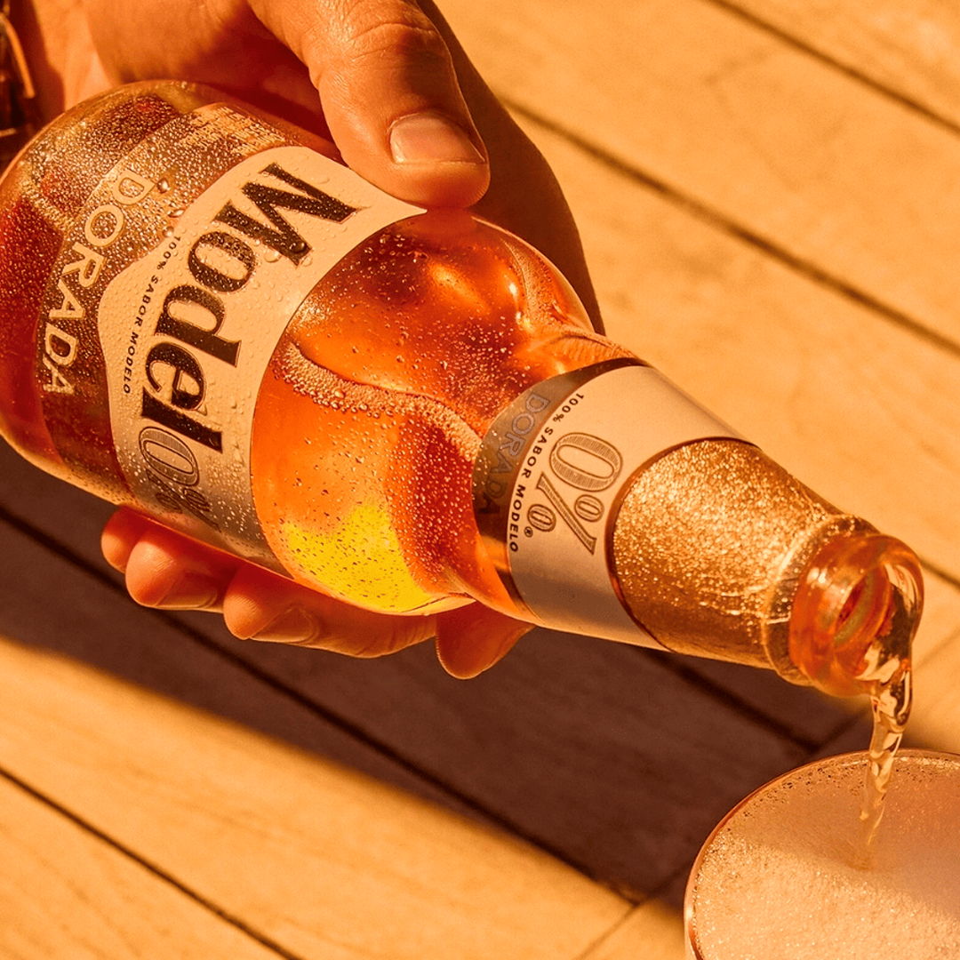 A hand pouring Modelo beer into a glass on a wooden surface.