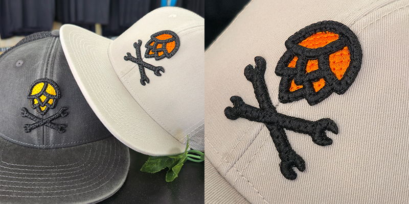 Close-up of brew garage logo featuring a beer hop and two crossed spanners, embroidered in quilted puff embroidery on grey and tan caps showing layered stitching with raised sections and detailed texture