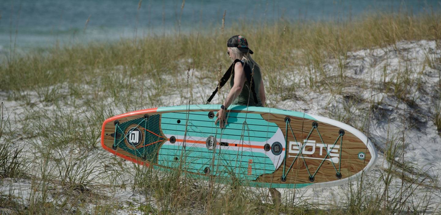 Woman carrying her Flood 10′6″ Classic Teak Paddle Board with Travelink Sling through sand dunes