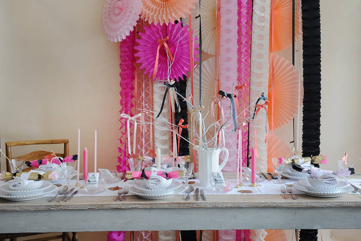 A table laid for a hen do with white pearl china, pink candles and a paper party decoration backdrop of fans and garlands in pinks, cream and black.