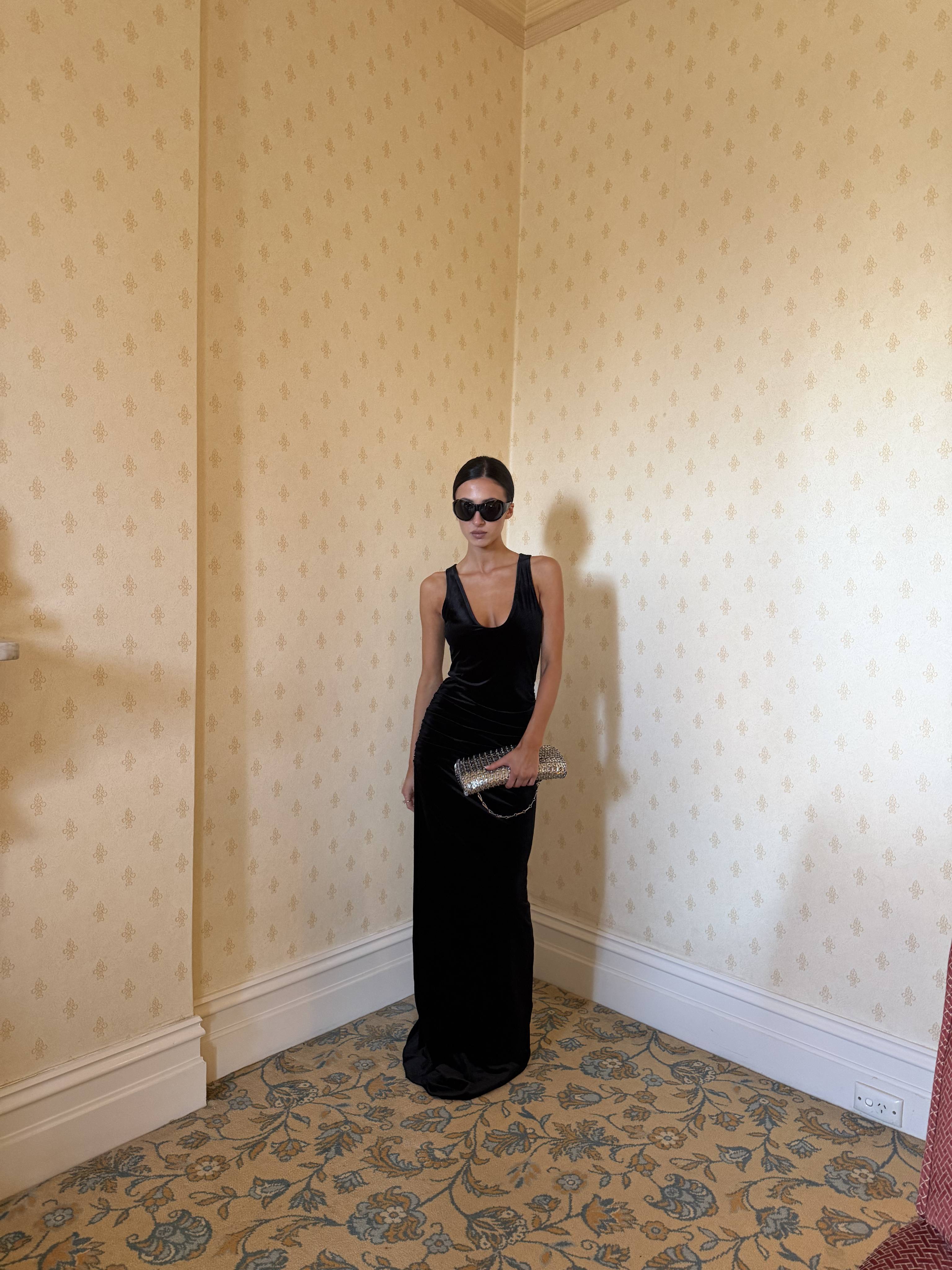 A person in a black dress and sunglasses stands in a corner.