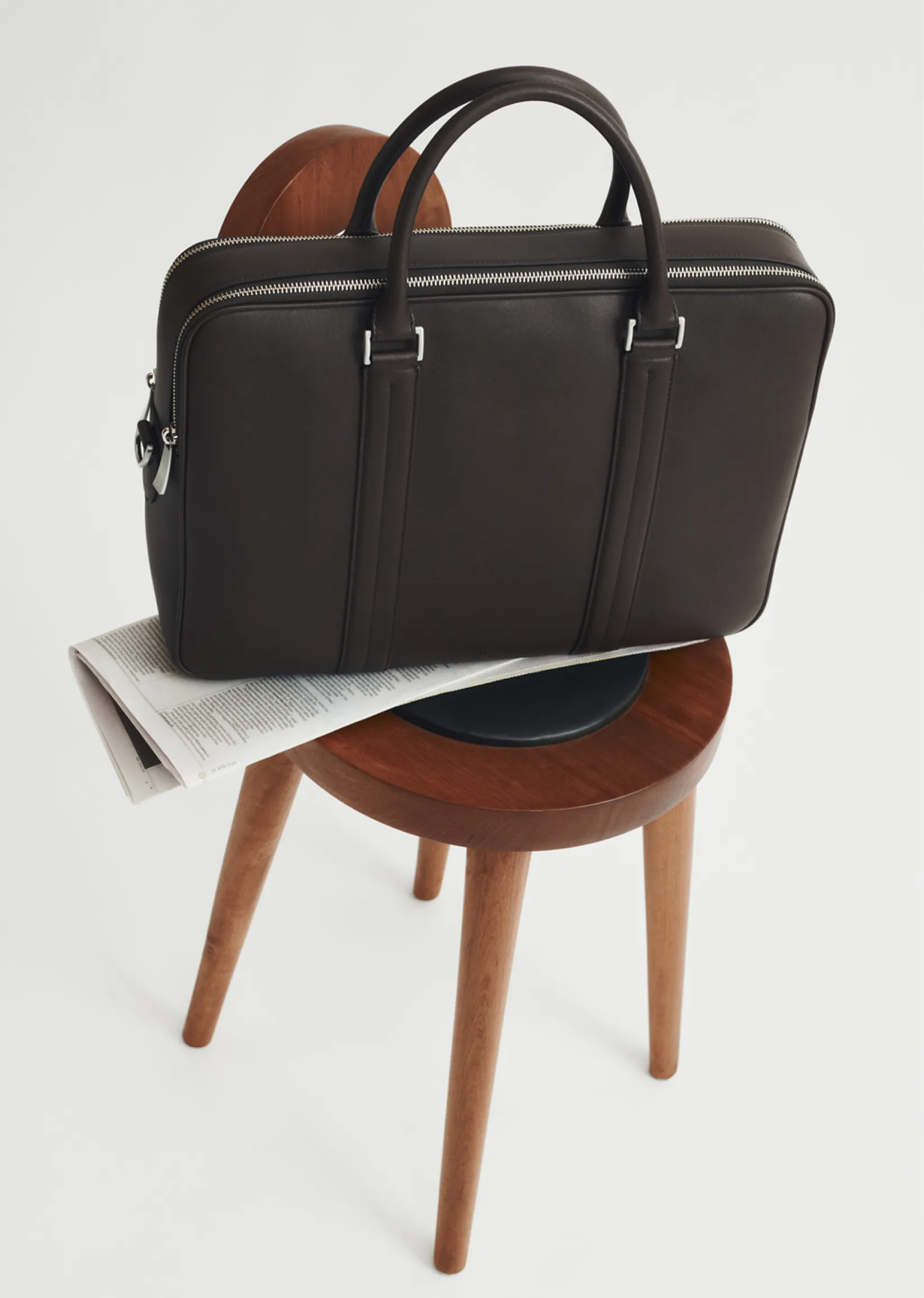 A brown leather bag on a wooden chair with a newspaper underneath.