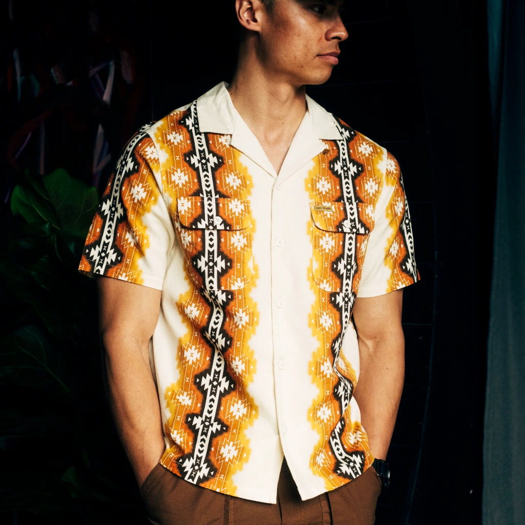 Man wearing a colorful patterned shirt, standing against a dark background.
