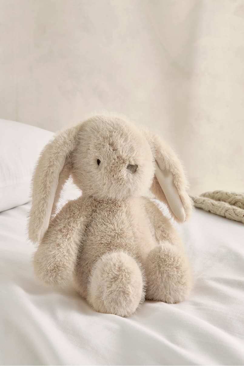 A soft, beige stuffed bunny sitting on a white bed.