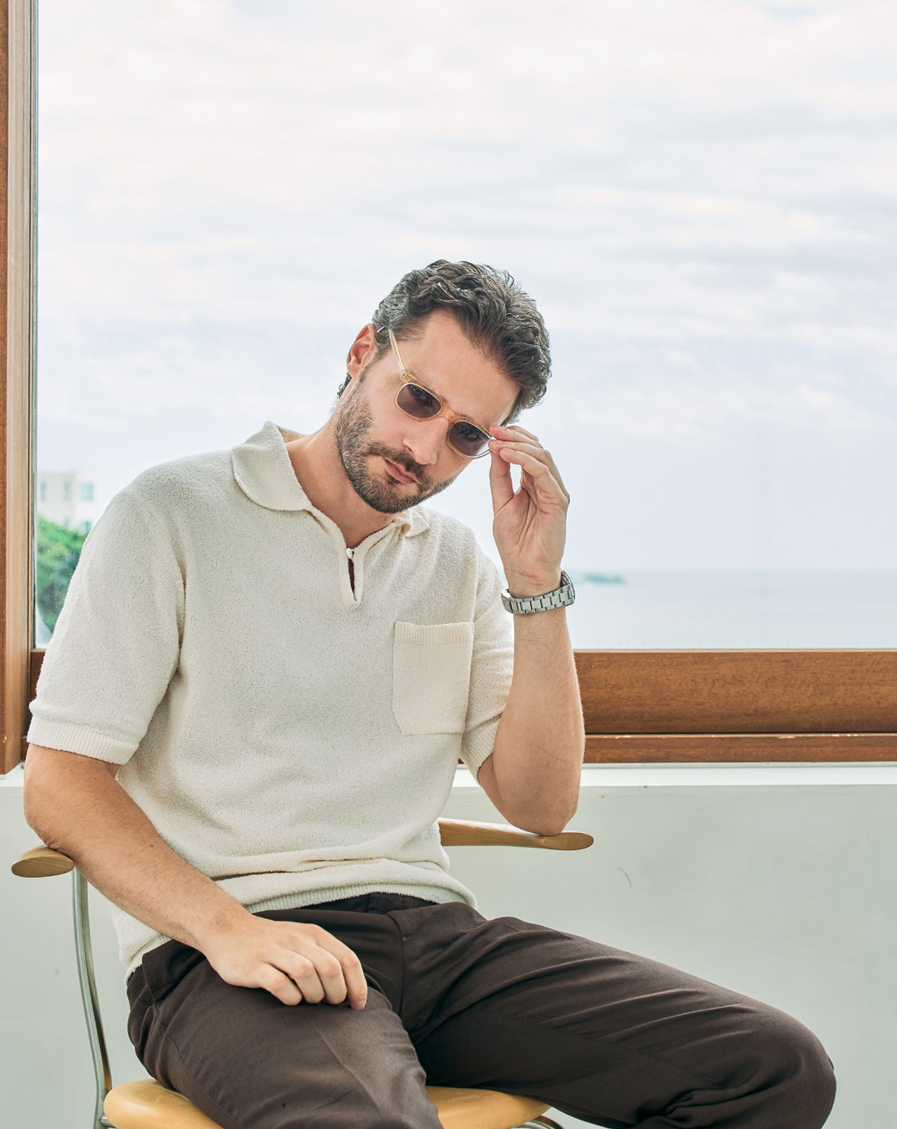 Man sitting on a chair, wearing sunglasses and light-colored clothing, looking thoughtful.