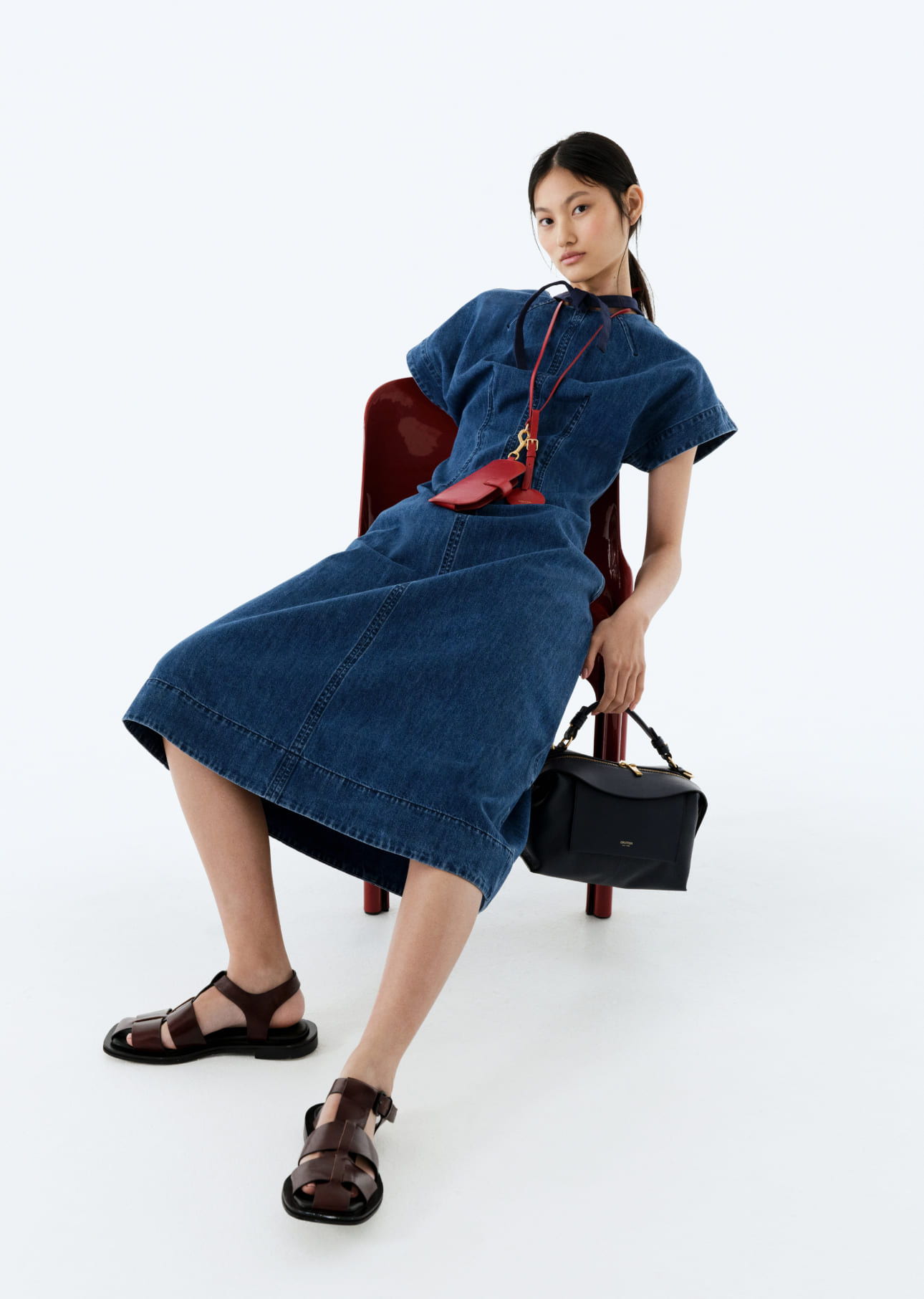 Model wearing a denim dress and sandals, seated on a chair.