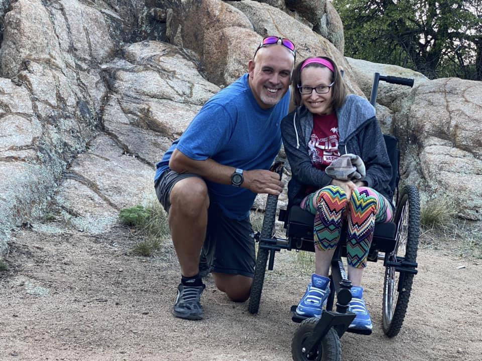 Patrick kneels next to Colleen smiling in GRIT Freedom Chair on dirt nature area with rocks behind them