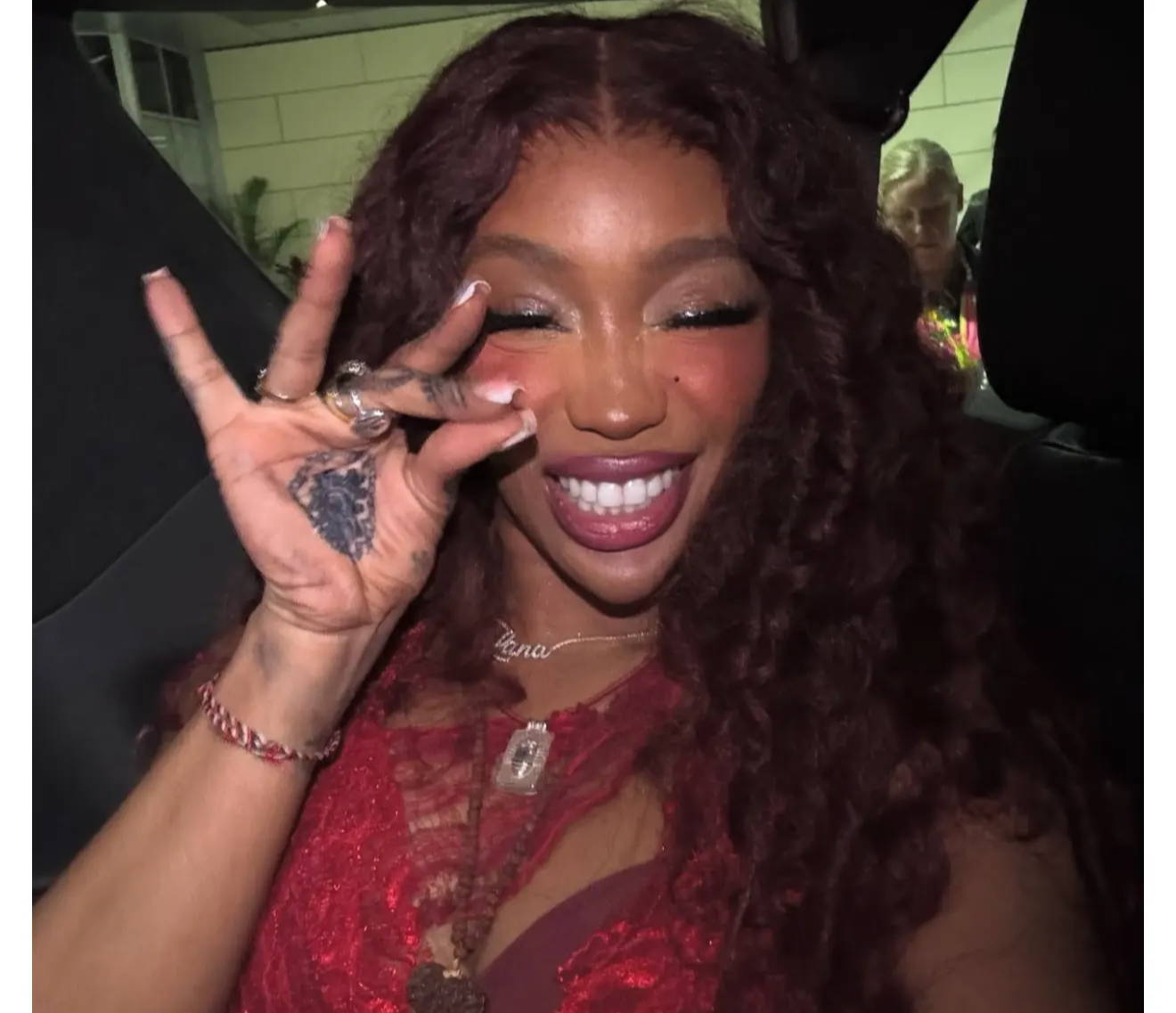 Smiling person with curly hair showing a hand gesture, wearing a red outfit.