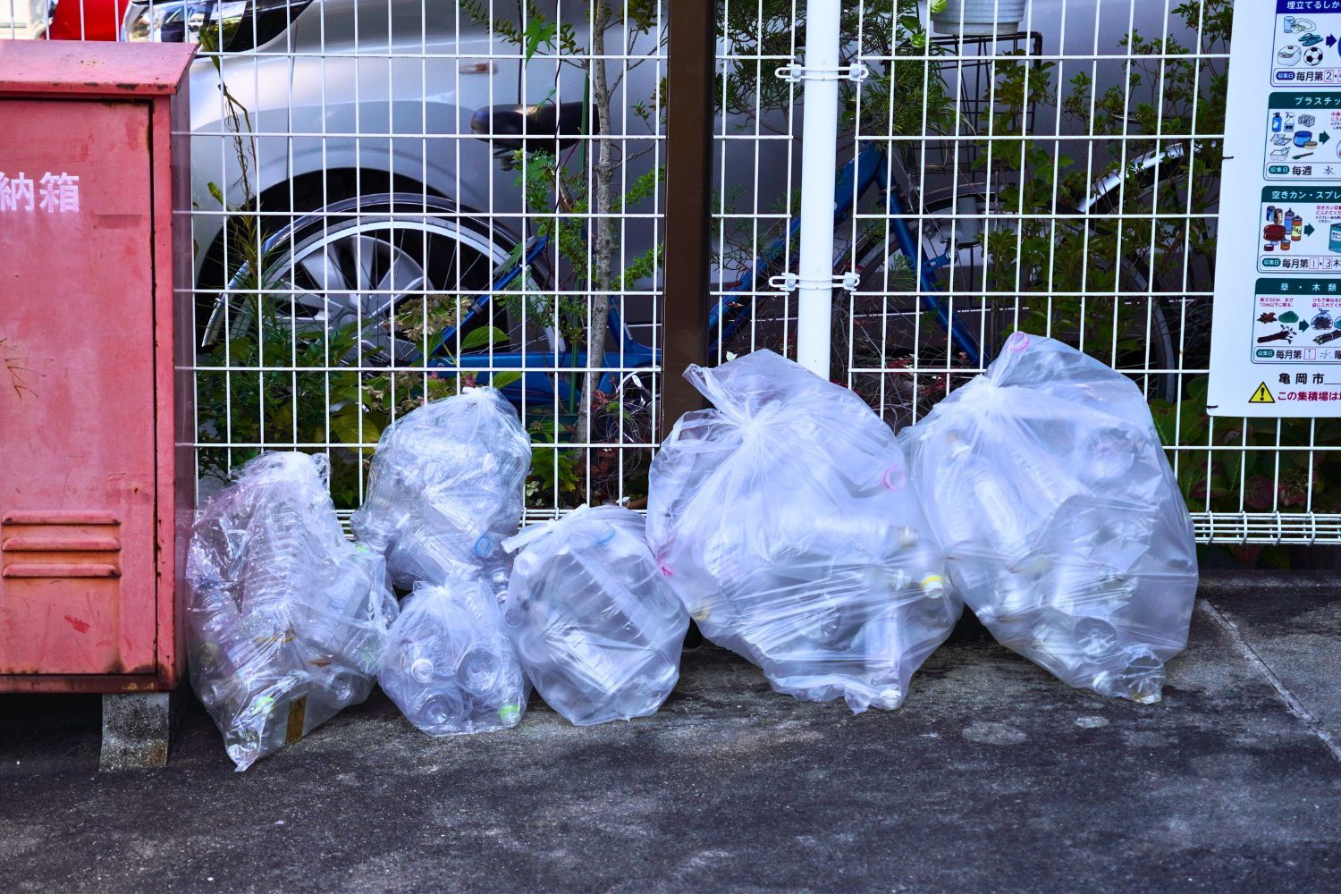 Several bags of waste near a fence and a dumpster.