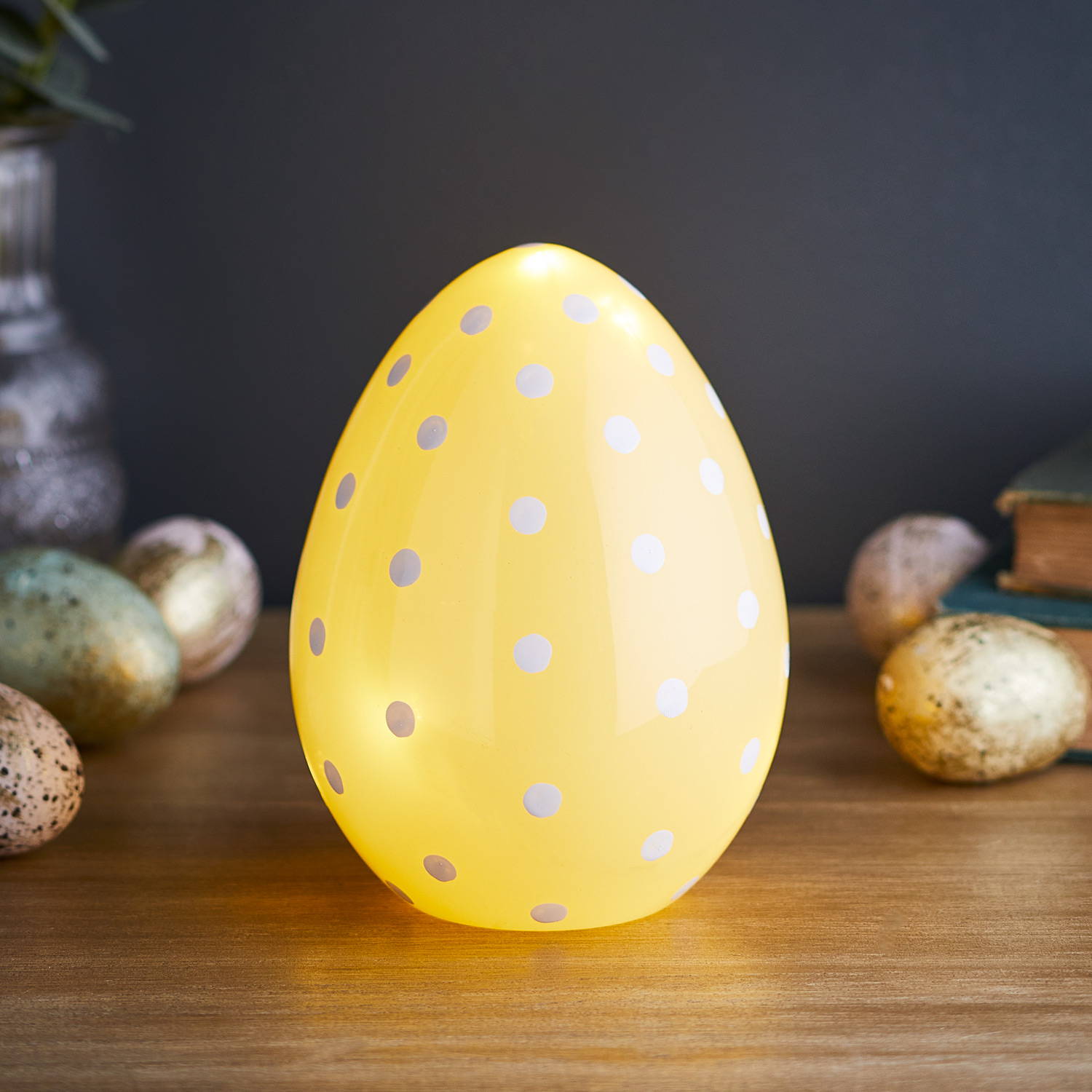 Yellow glass Easter egg light illuminated on table with chocolate eggs scattered next to it