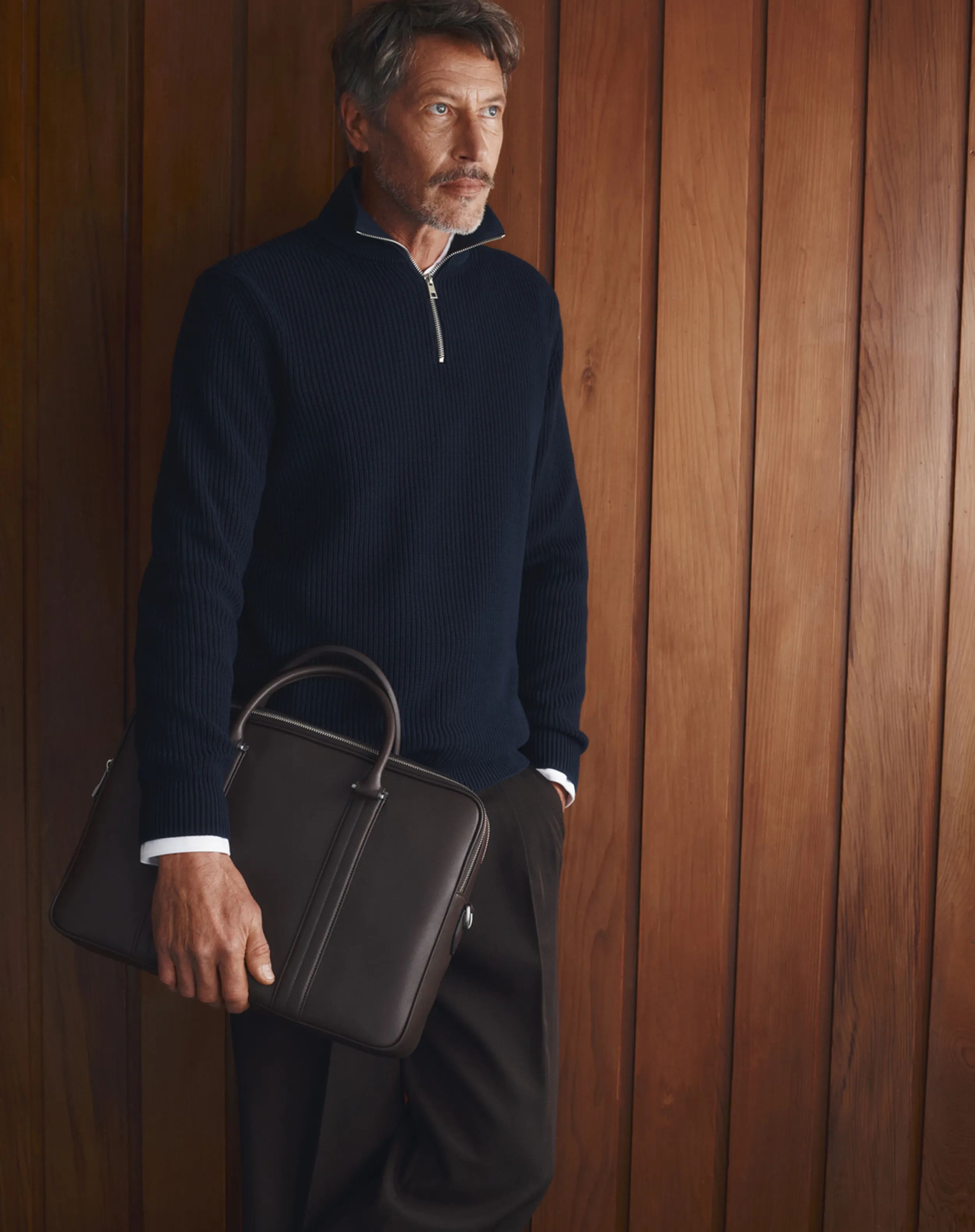 Man in a navy sweater holding a brown leather bag against a wooden wall.