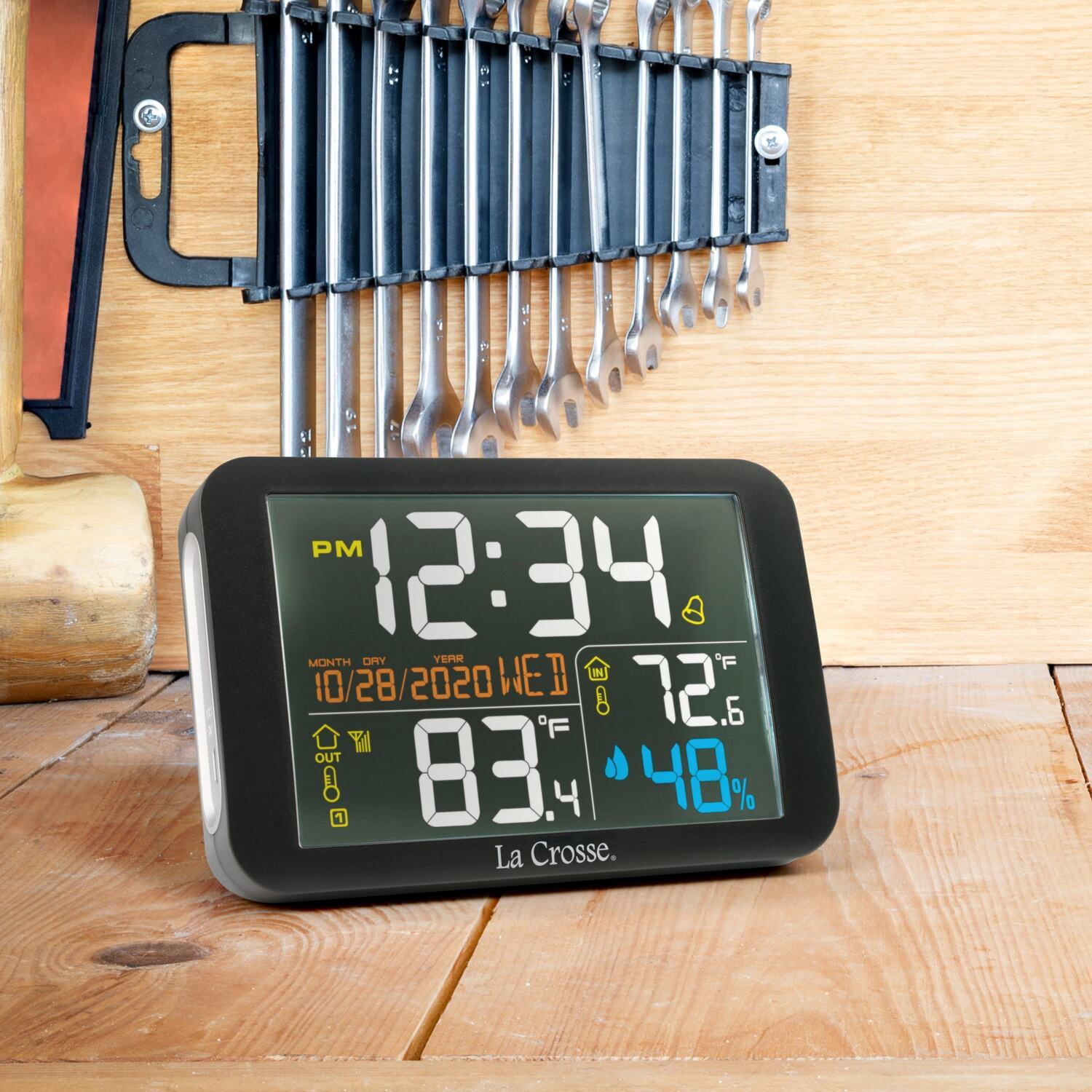 Digital clock and weather station on a wooden surface with tools in background.