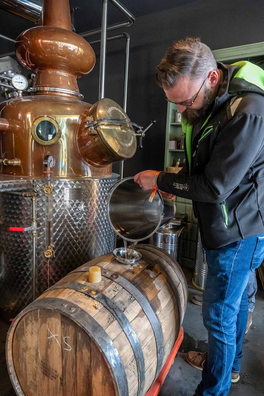 Person pouring liquid from a pot into a wooden barrel near a distillery.