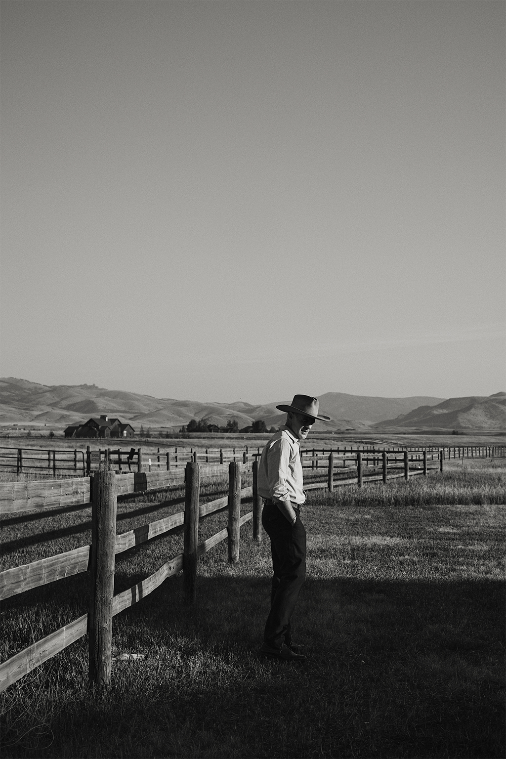 A black and white photograph of Michael Kelly standing next to a fence in Pray, Montana wearing a vintage stetson, the Rivay Bridges Oxford Workshirt in Navy, Rivay Italian Wool Suit in Navy Chalk Stripe Trousers and Rivay X Alden Bedford Boot in Shell Cordovan Color 8 