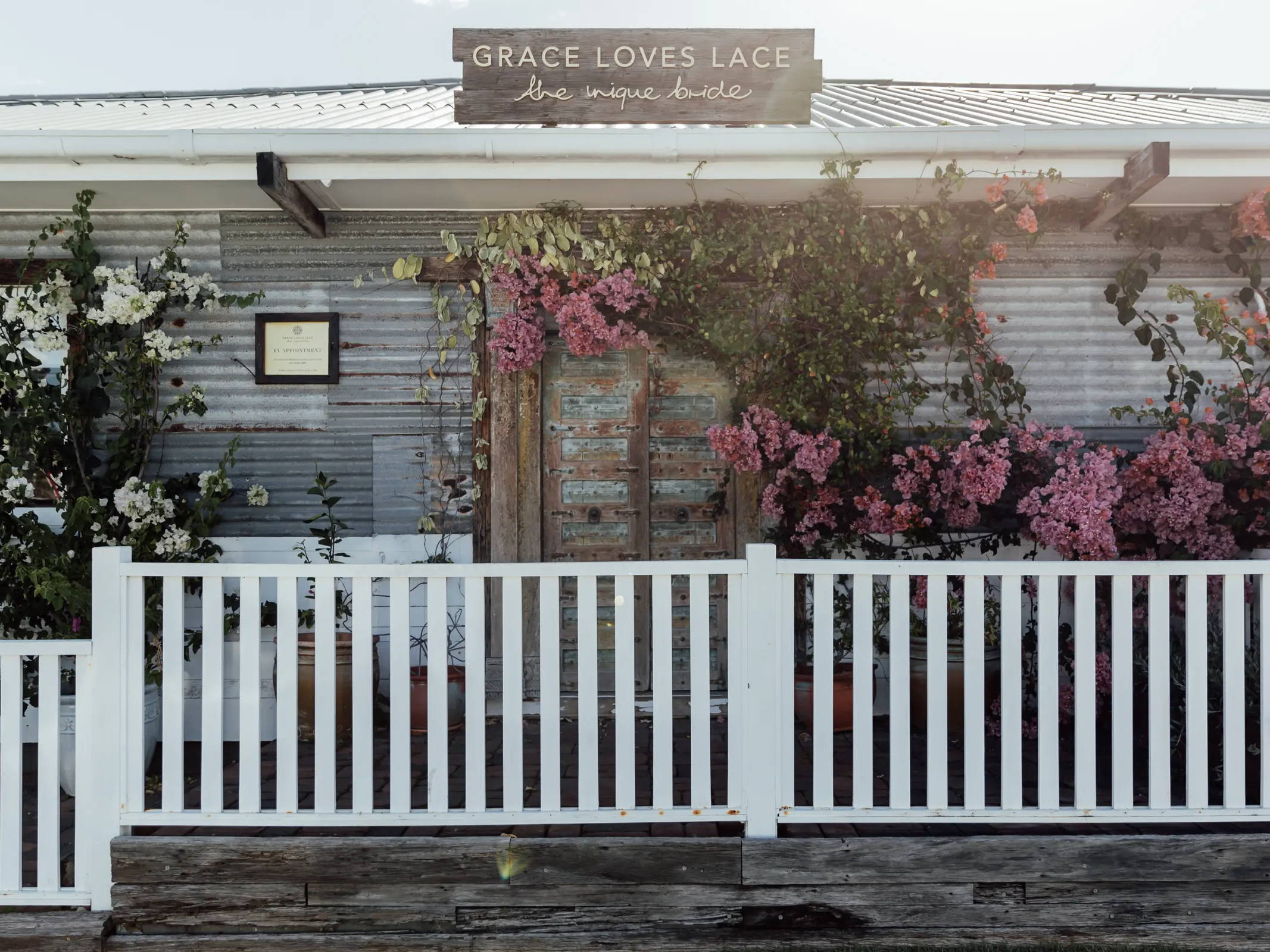El exterior de la sala de exposición de Gold Coast Grace Loves Lace con valla blanca y buganvilla rosa.