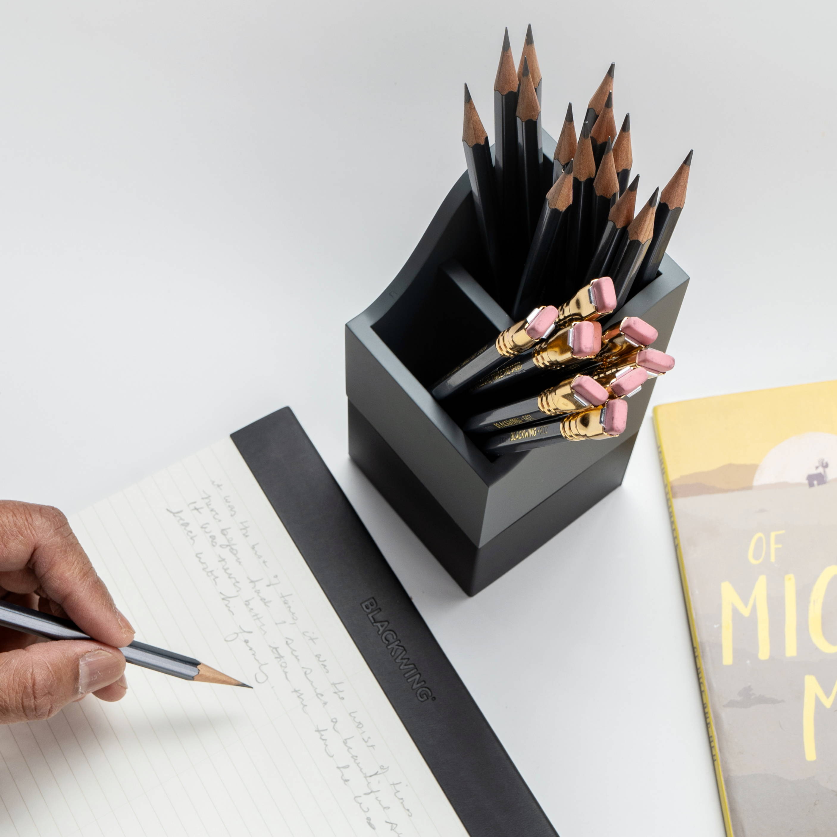 A hand writing on a notepad beside a pencil holder and a book.