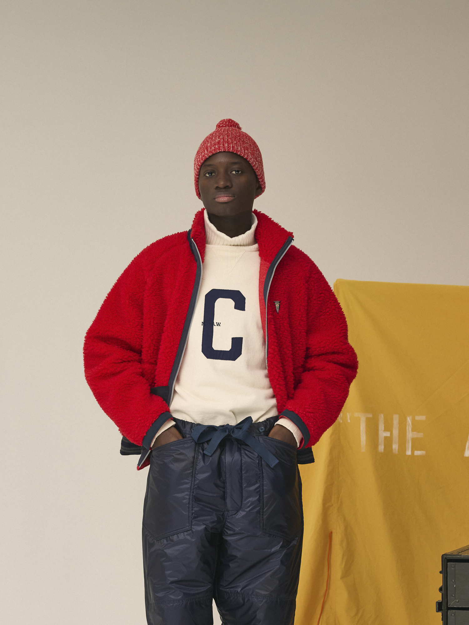 Person wearing red jacket and hat, standing beside yellow backdrop and storage boxes.