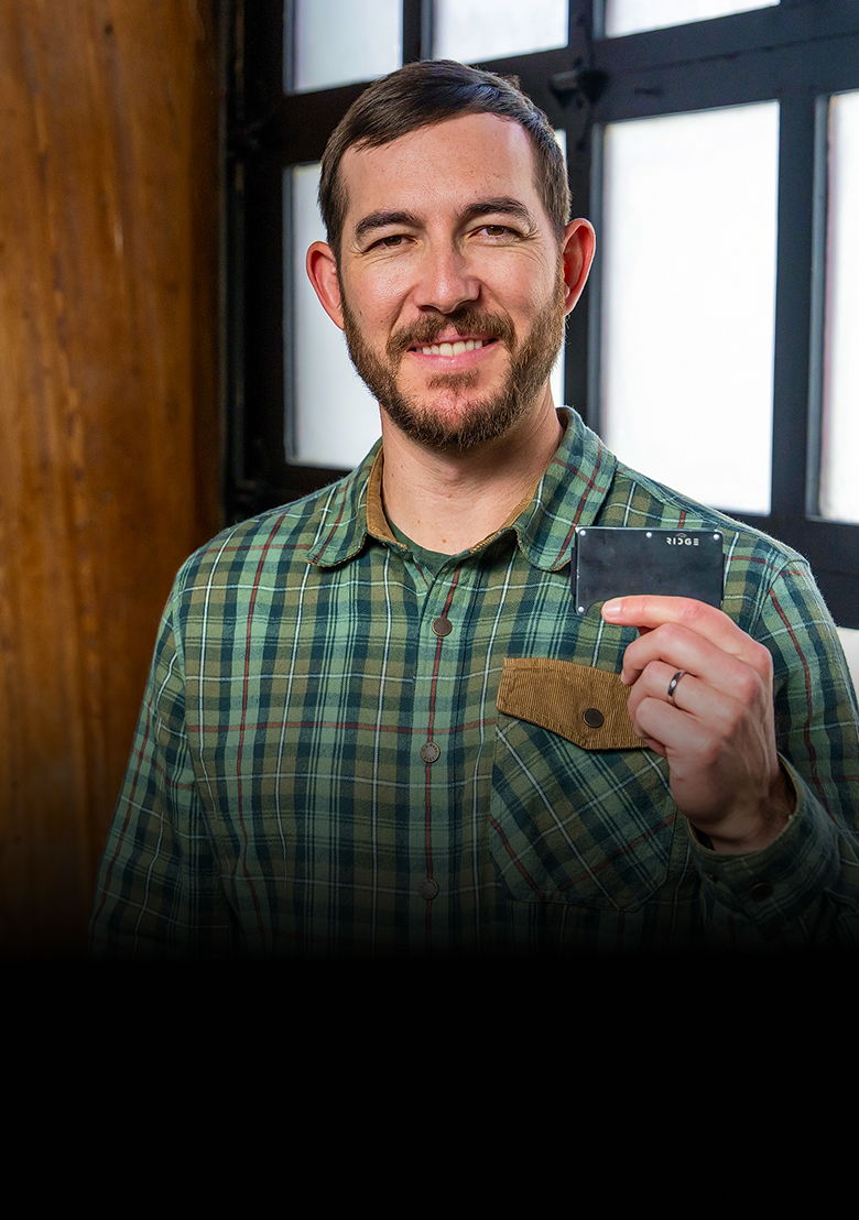 Man in a plaid shirt holding a card, smiling in front of a window.