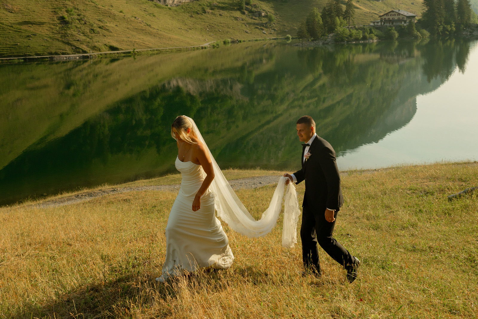 A couple in wedding attire walking by a serene lake.