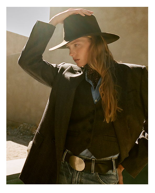 Person wearing a black hat and long coat, striking a thoughtful pose outdoors.