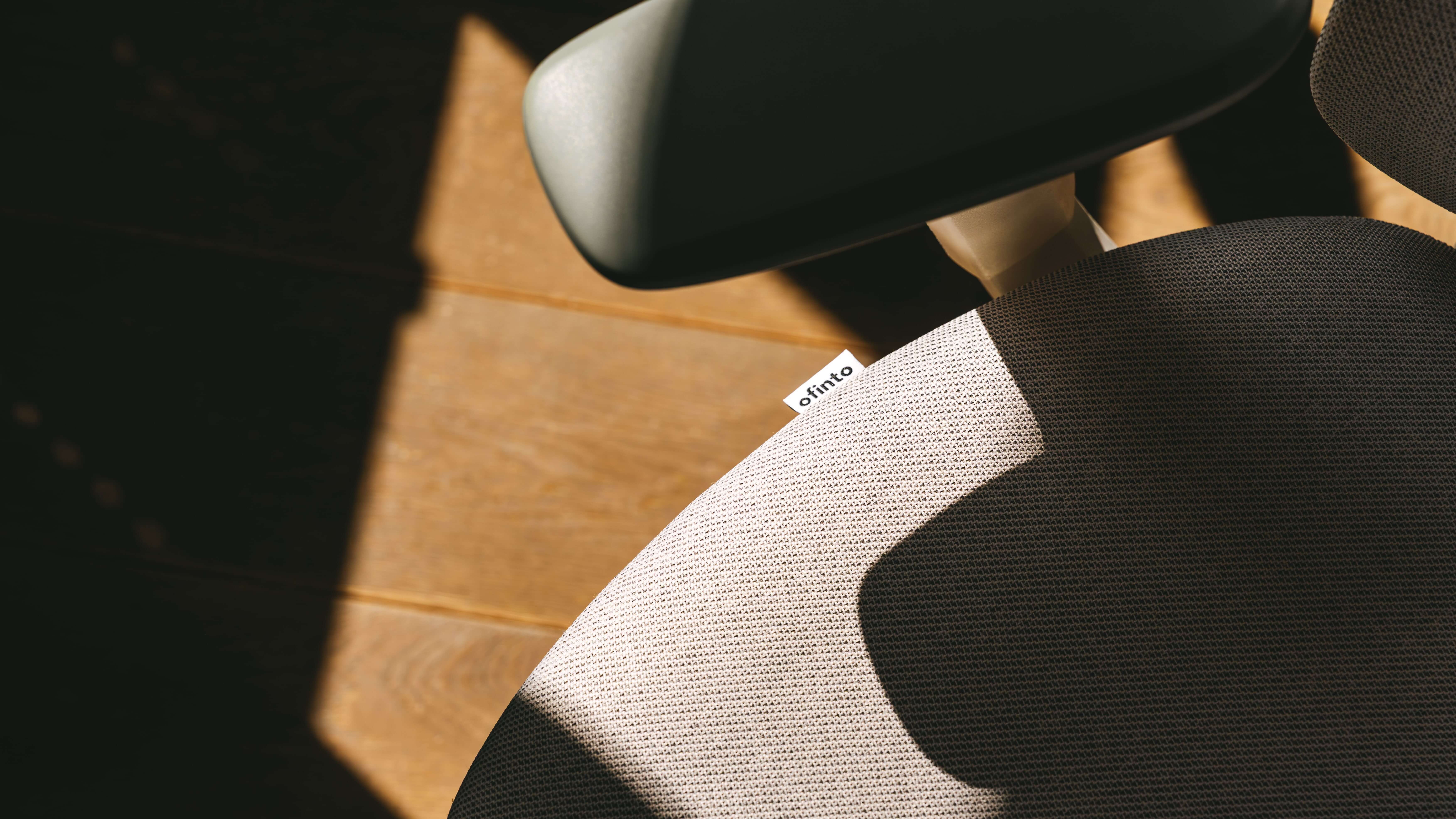 Detailaufnahme eines hellen Bürostuhlsitzes mit „ofinto“-Label, schwarzer Armlehne und Holzboden, Licht und Schatten im Bild.