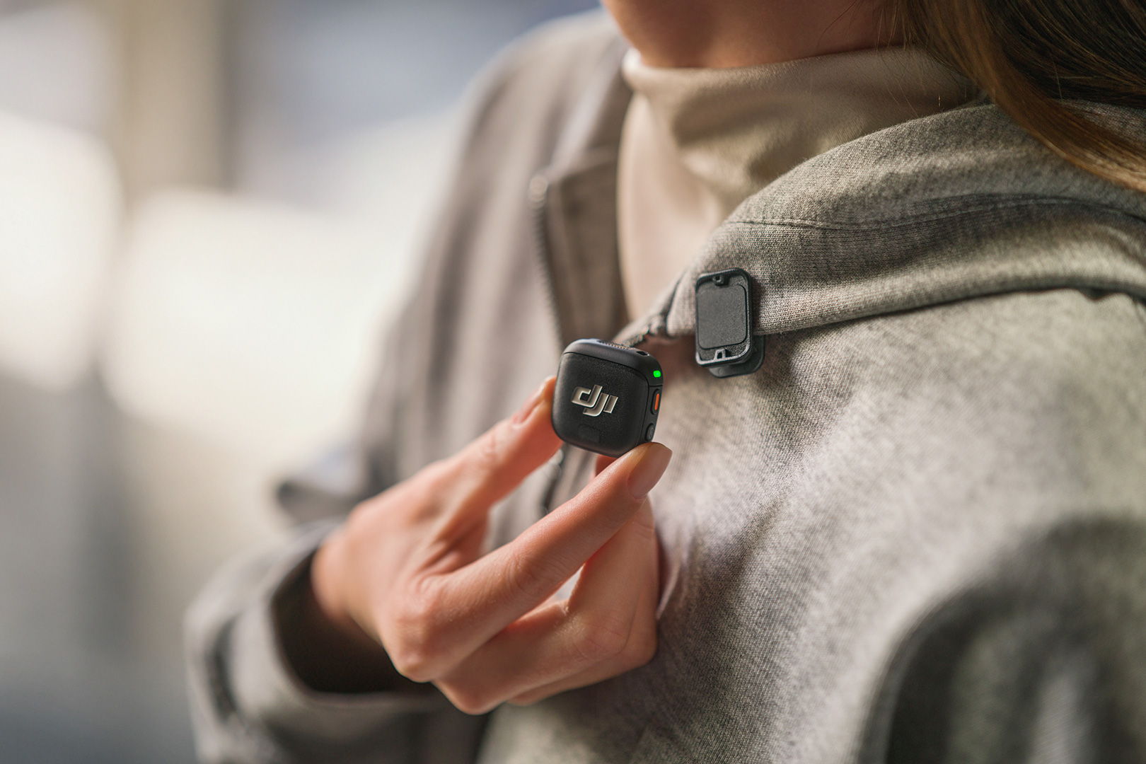 Person adjusting a DJI Mic 3 attached to a gray hoodie.