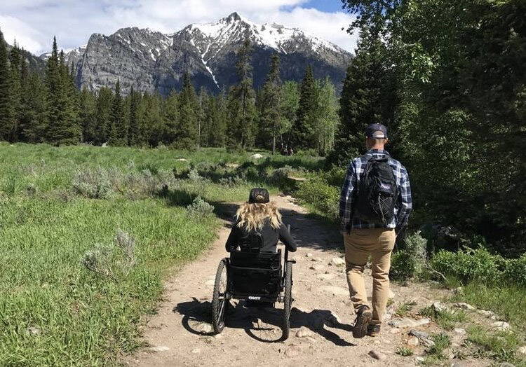 Pair hiking, one using all terrain GRIT Freedom Chair wheelchair, on rocky dirt path through grass and forested mountain area