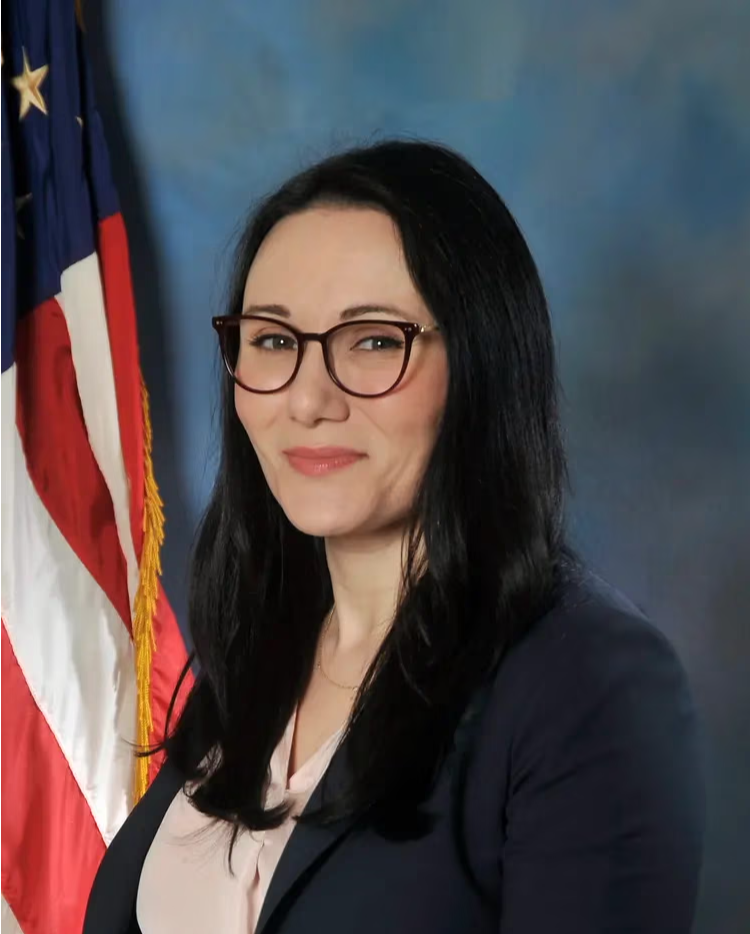 A woman in a suit smiling, with an American flag in the background.
