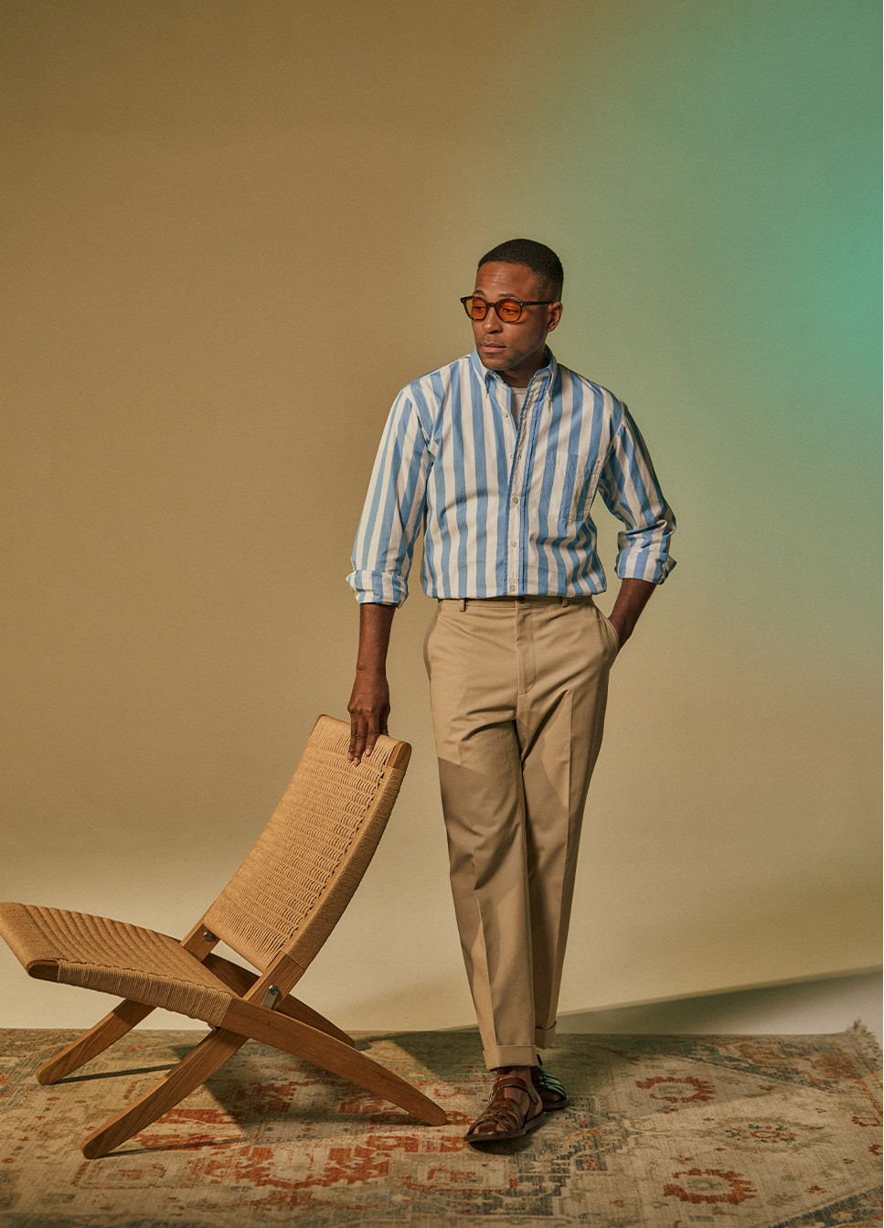 Man in striped shirt and khakis poses next to a wooden chair.