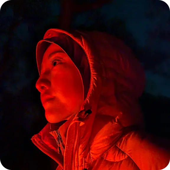 Person in a hooded jacket illuminated by warm light against a dark background.