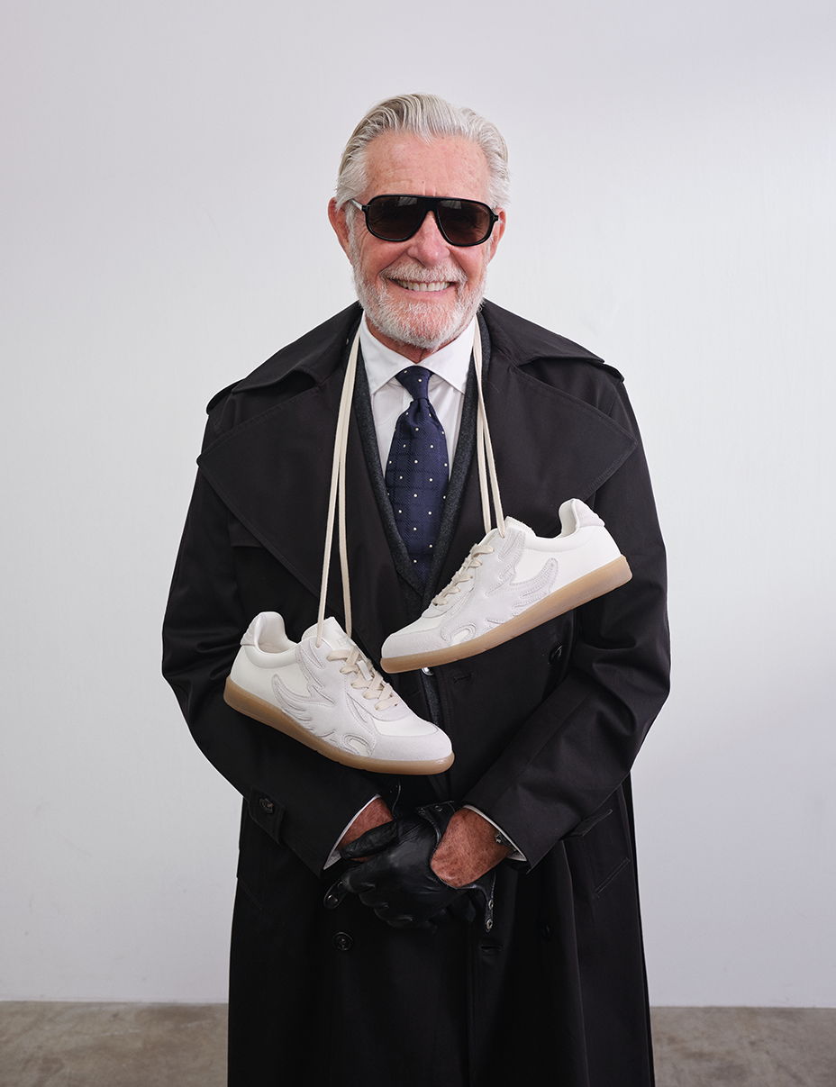 Older man in sunglasses and coat holding white sneakers.