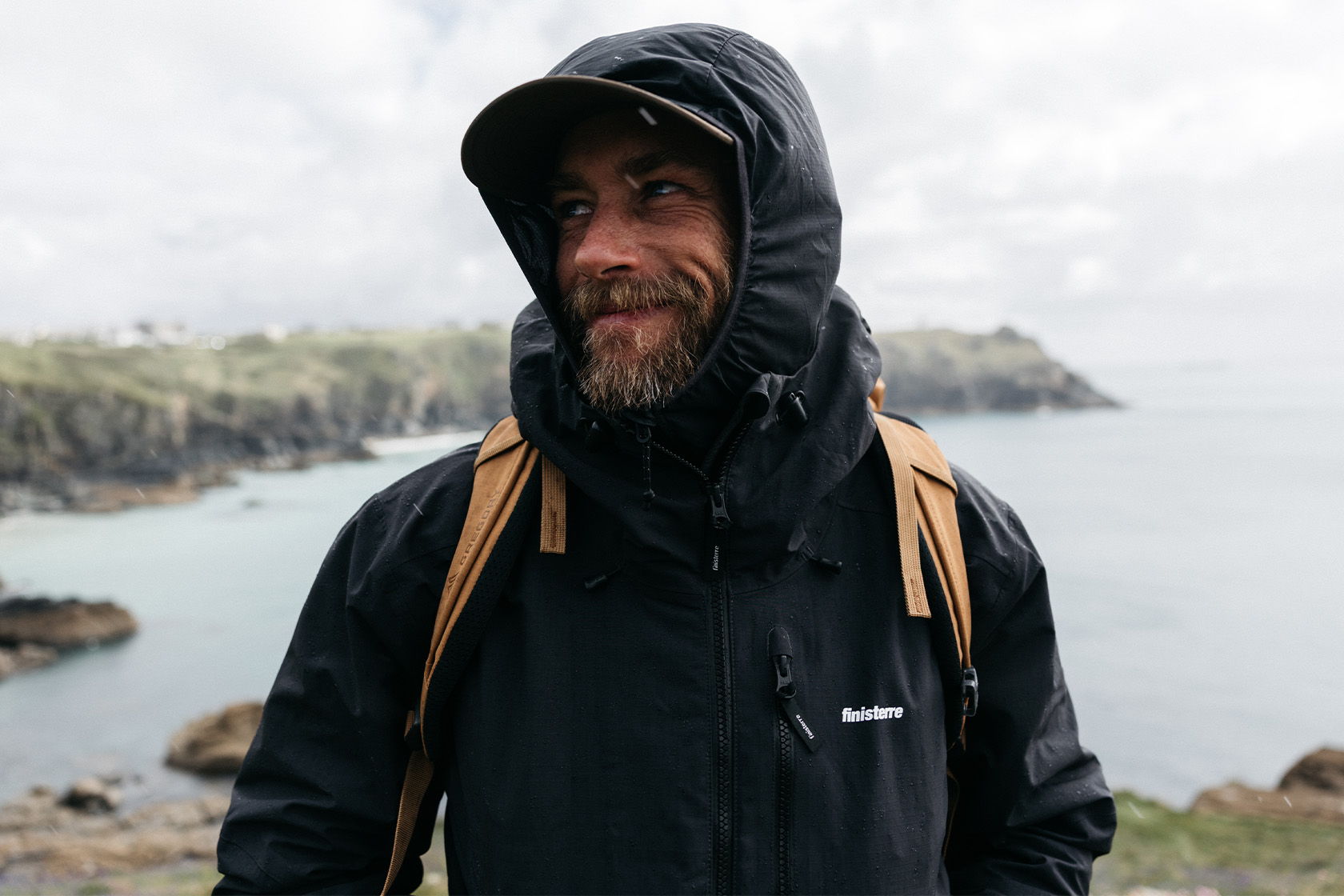 Person smiling in a jacket by the sea with rocky landscape.