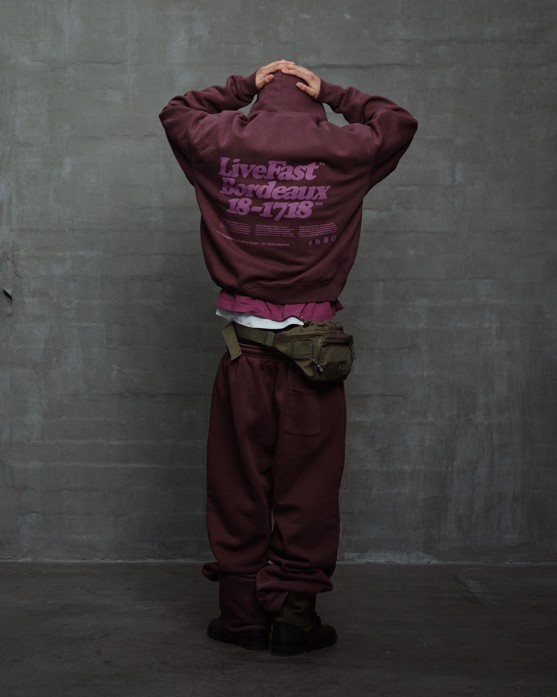 Person in maroon outfit facing away, hands on head, against gray background.