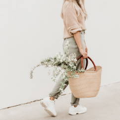 woman walking with green pants, tan shirt, white tennis shoes, with wicker basket and white flowers