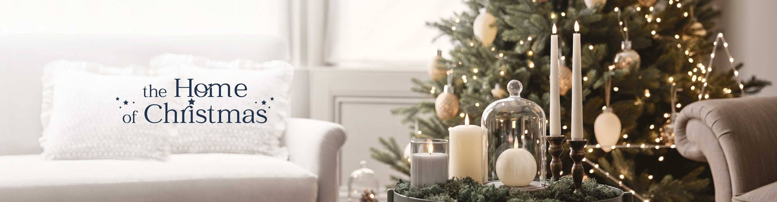 Verschiedene TruGlow LED Kerzen und eine Glasglocke auf einem Beistelltisch im Wohnzimmer, dahinter ein beleuchteter Weihnachtsbaum