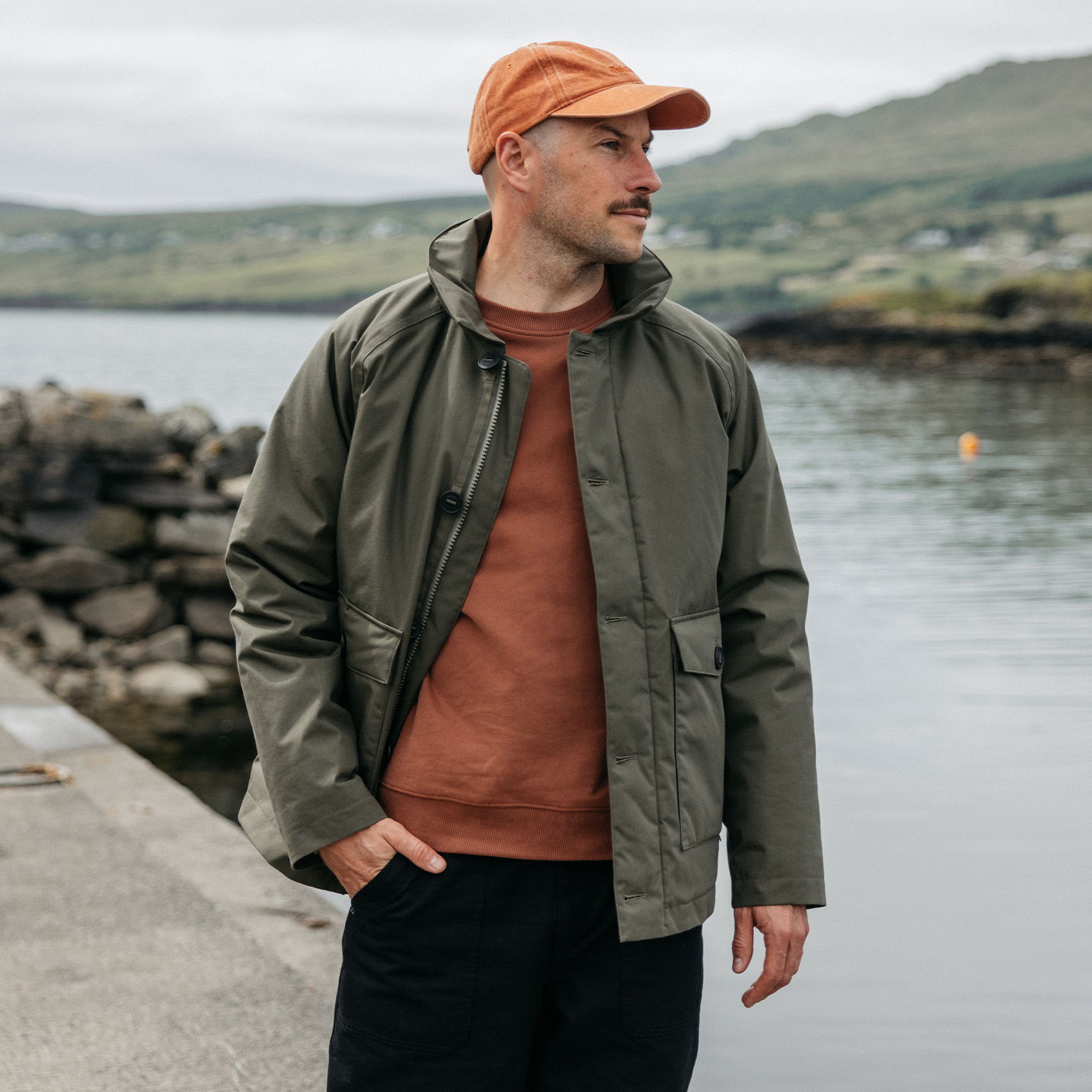 Man in an olive jacket and orange cap by the water.
