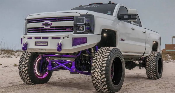 Front three-quarter view of Ash's white lifted 2015 Chevrolet Silverado 2500HD with purple suspension and large off-road wheels on the beach, Conductor's Special 244K install photo 1