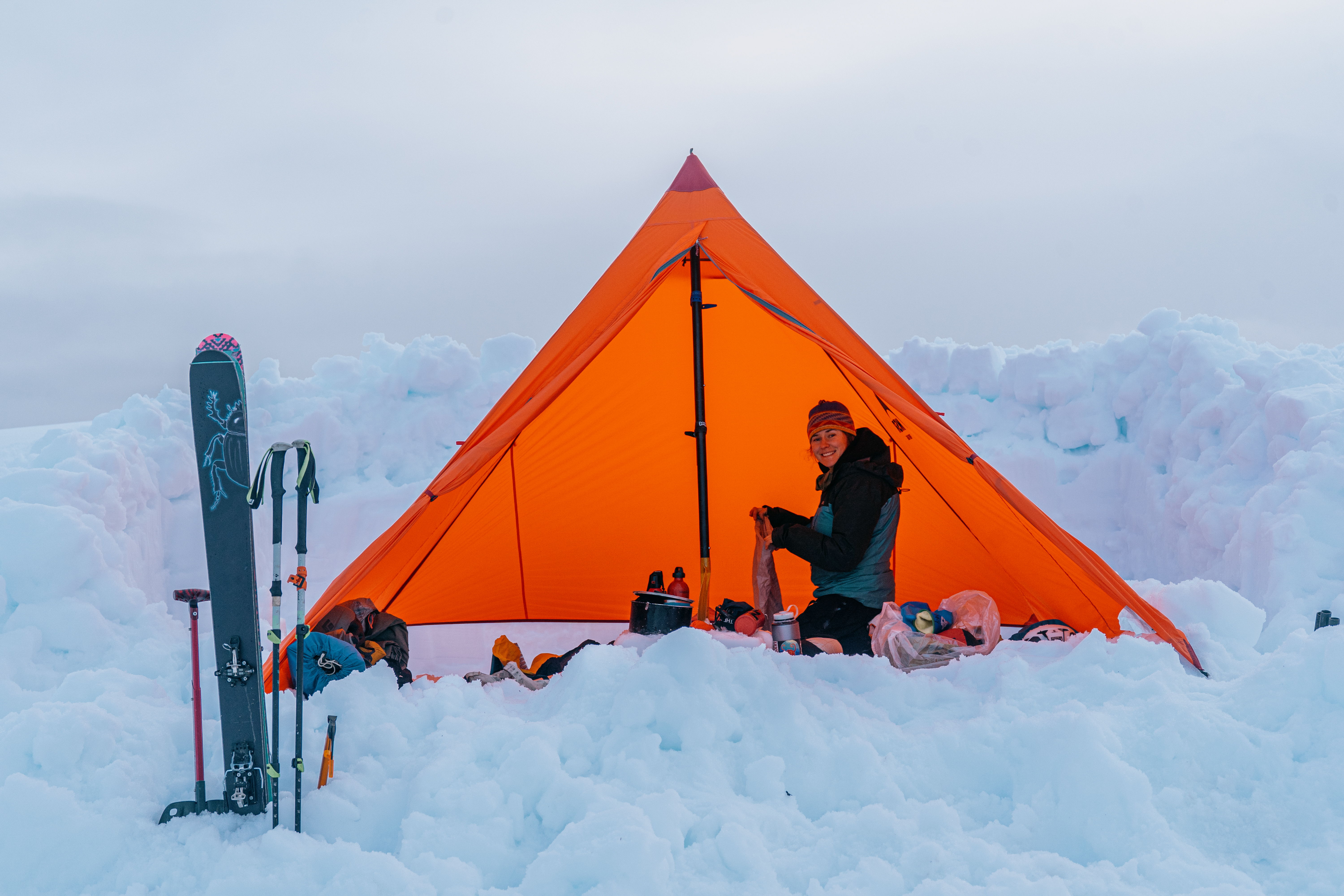 Person sitting by an orange tent in a snowy landscape with splitboarding equipment.