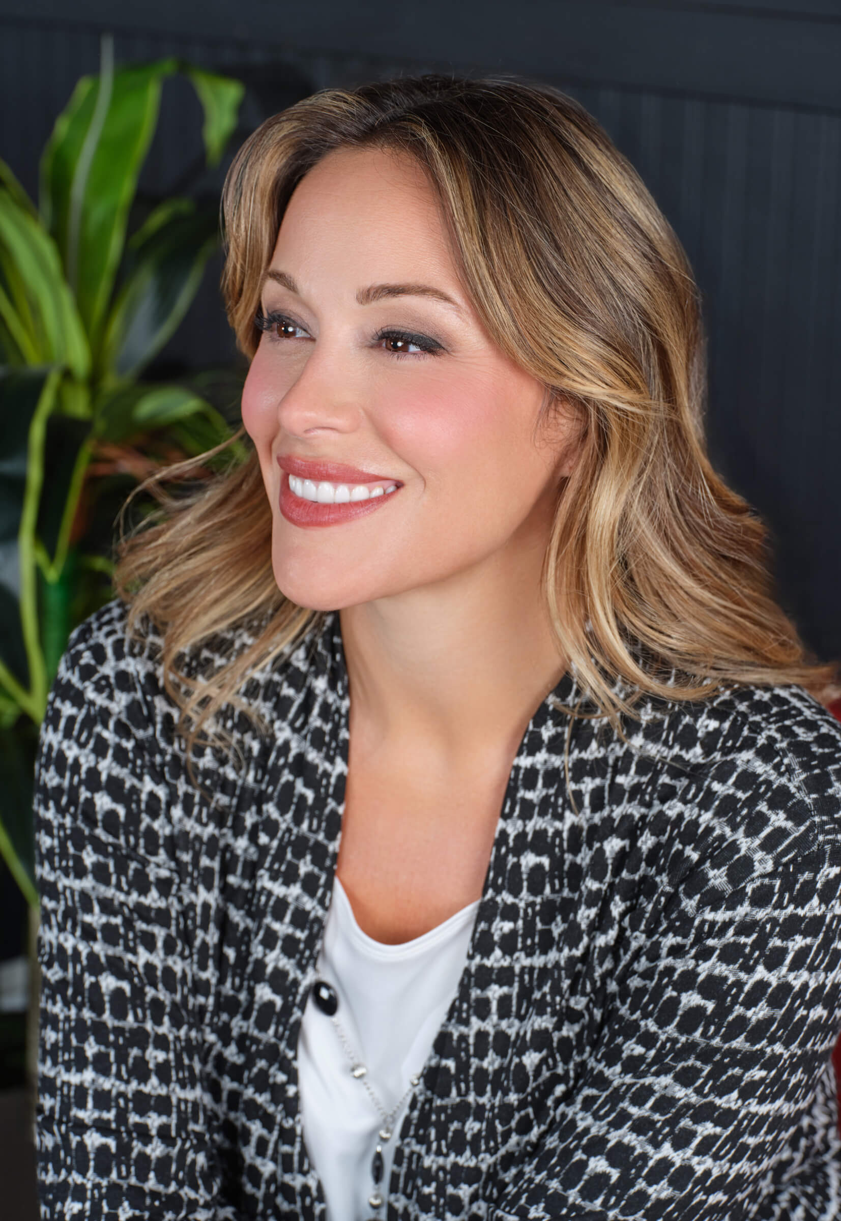 Smiling woman in a patterned jacket with a plant in the background.
