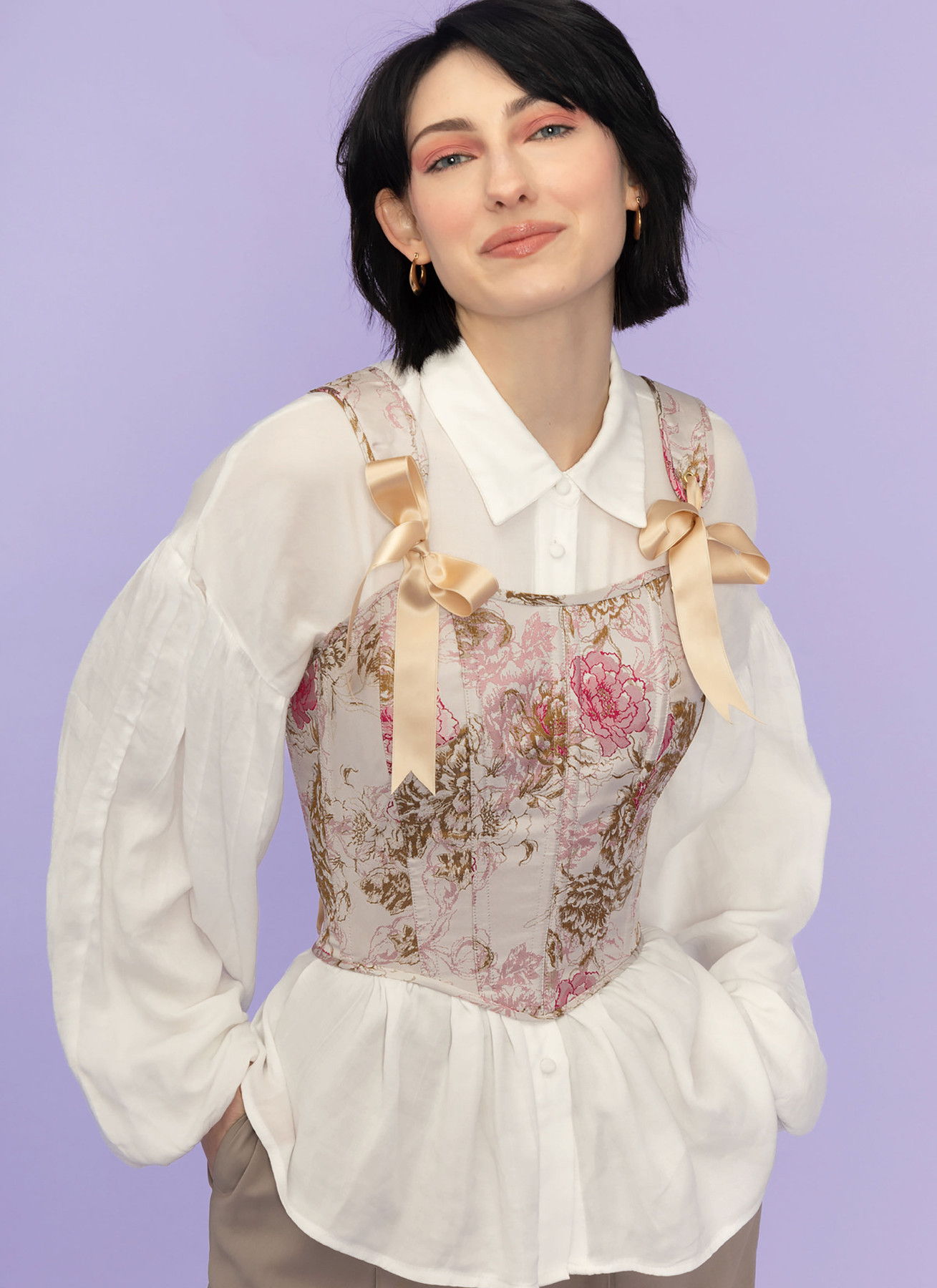 Person wearing a floral corset over a white blouse, smiling against a purple background.