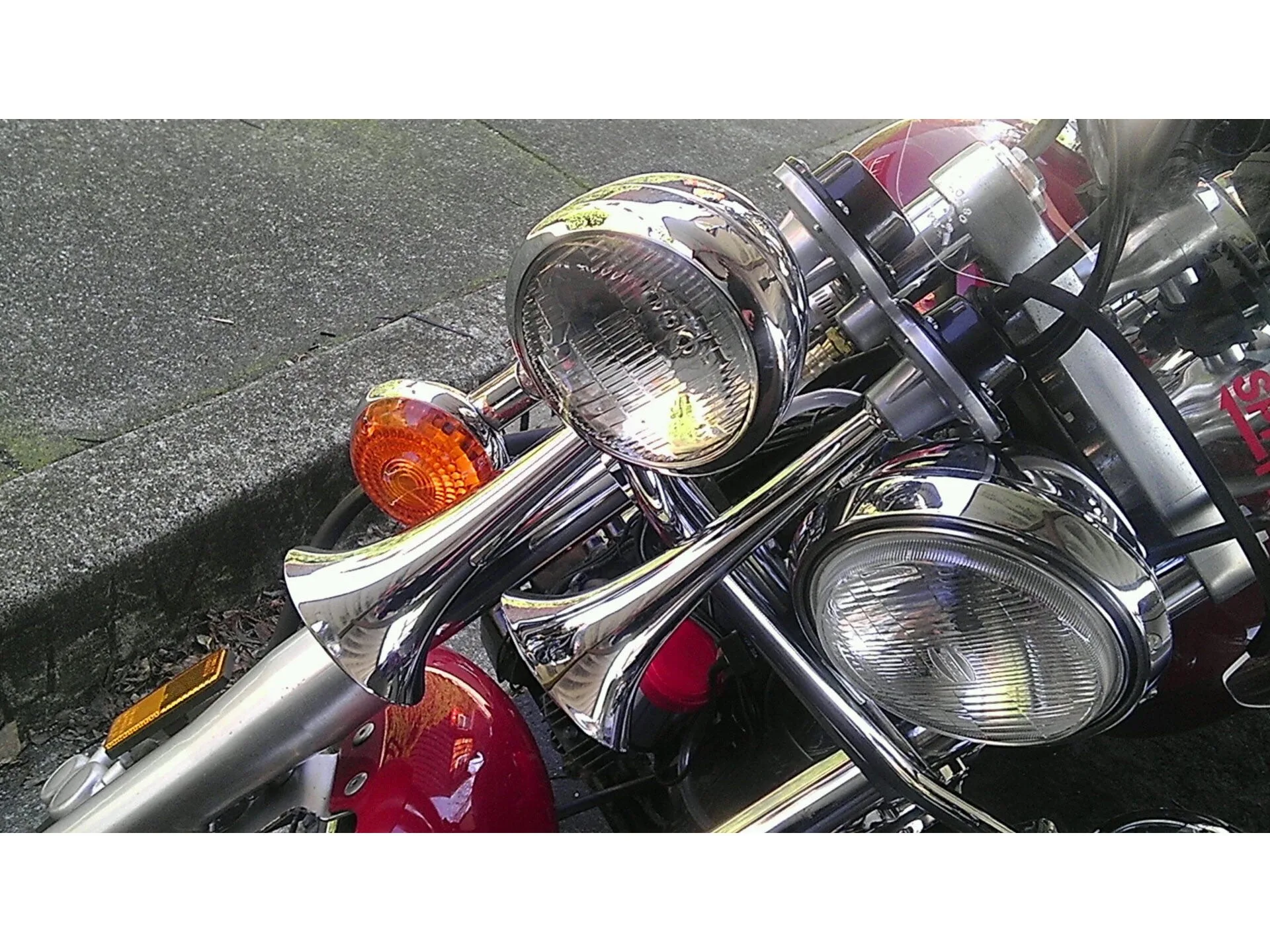 High-angle close-up Ty's red Yamaha V-Star cruiser front forks chrome PsychoBlasters V2 dual trumpet horns bracket wires mounted beside round headlight amber turn signal red front fender asphalt curb gallery photo 1