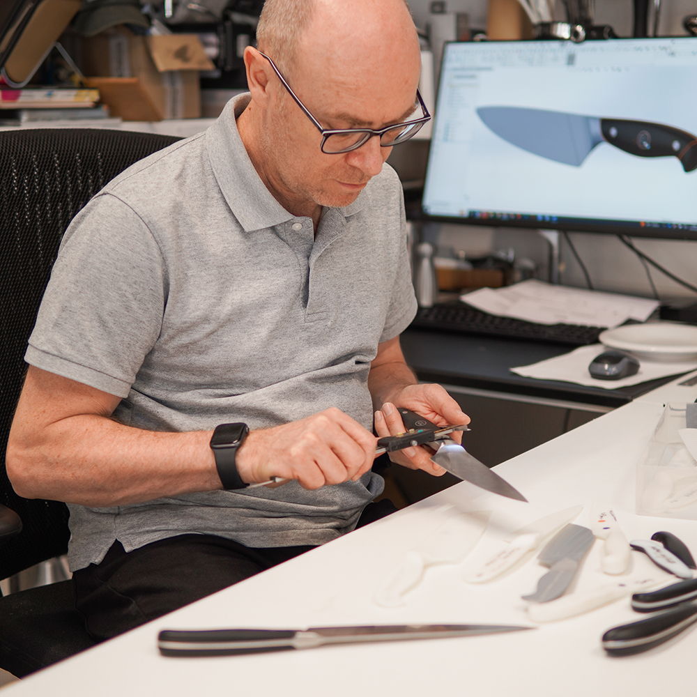 Paul working on knife design at a desk with tools and computer.