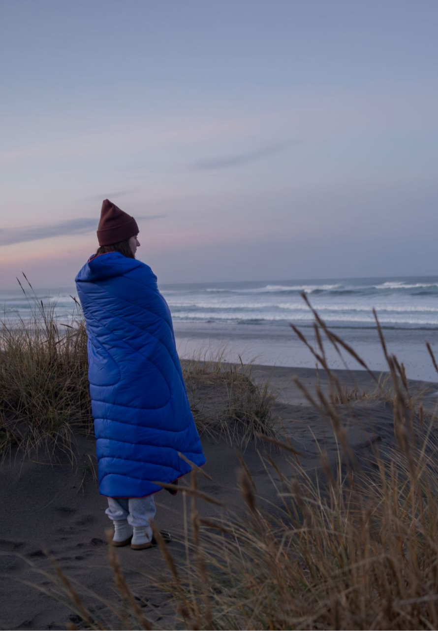 Person in a blue blanket standing on a beach at sunset.
