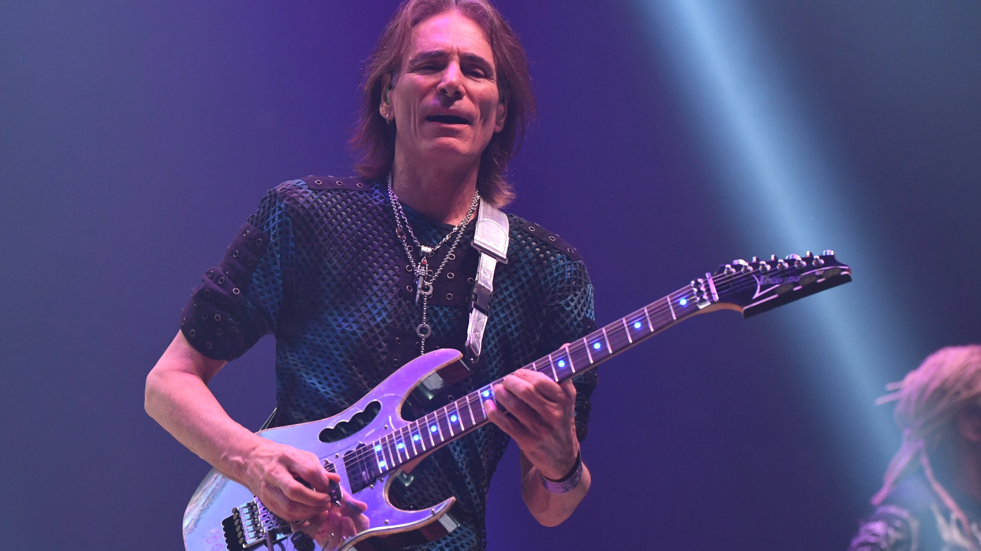 Steve Vai playing electric guitar on stage with dramatic lighting.
